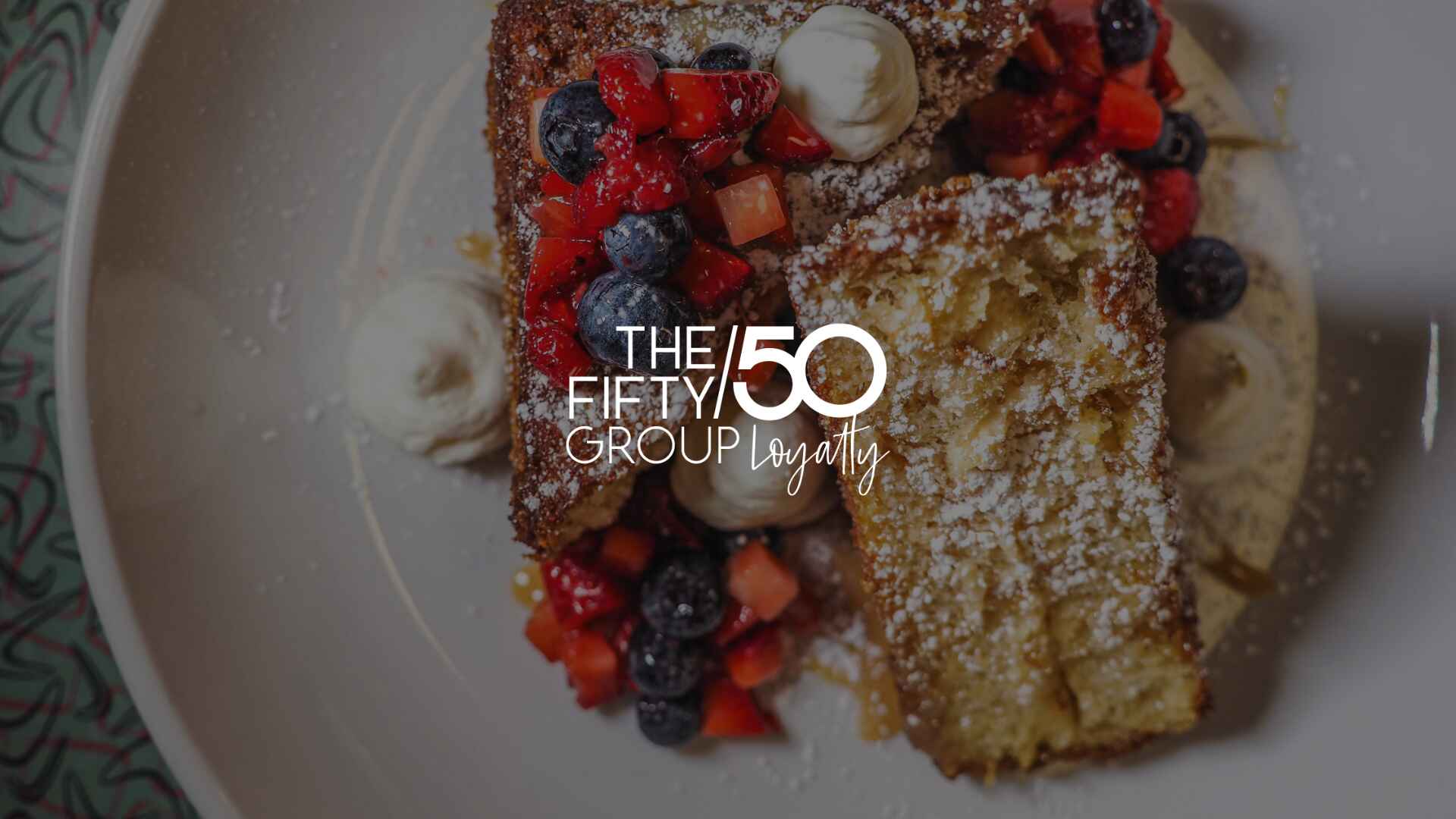 Plated dessert featuring French toast topped with fresh strawberries, blueberries, and whipped cream, branded with